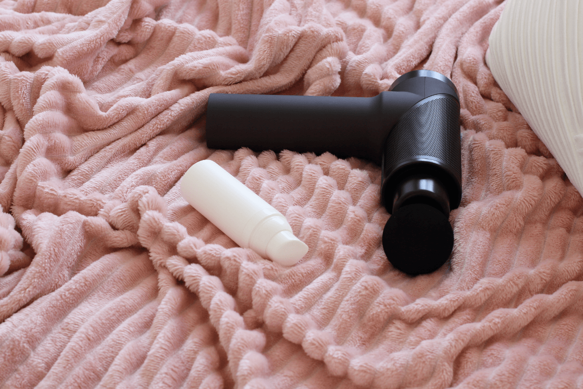 Pistola massageadora de alta vibração preta sobre manta rosa com frasco de óleo, ideal para massagem relaxante e estímulo corporal potente.