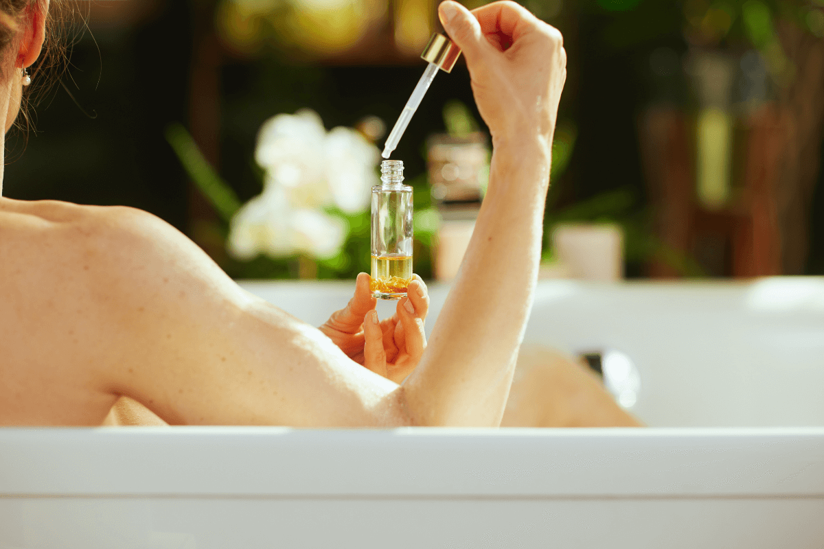 Mulher relaxando na banheira segurando um frasco de vidro com conta-gotas contendo óleo dourado, pronta para aplicar na água.