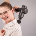 Mulher demonstrando como usar pistola de massagem nas costas e ombros corretamente para aliviar a tensão muscular sem ajuda profissional.