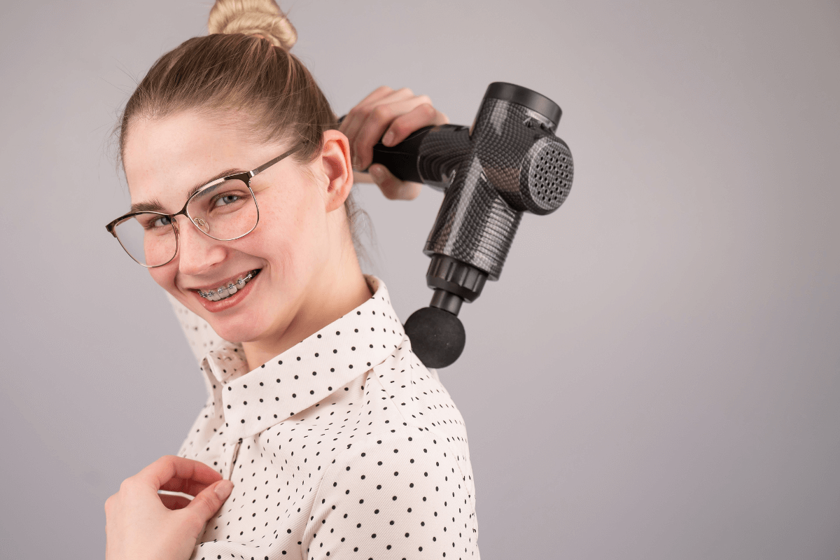 Mulher demonstrando como usar pistola de massagem nas costas e ombros corretamente para aliviar a tensão muscular sem ajuda profissional.