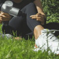 Mulher sentada no gramado demonstrando como usar a pistola massageadora na perna, aplicando o aparelho prateado na região lateral da panturrilha para recuperação muscular.