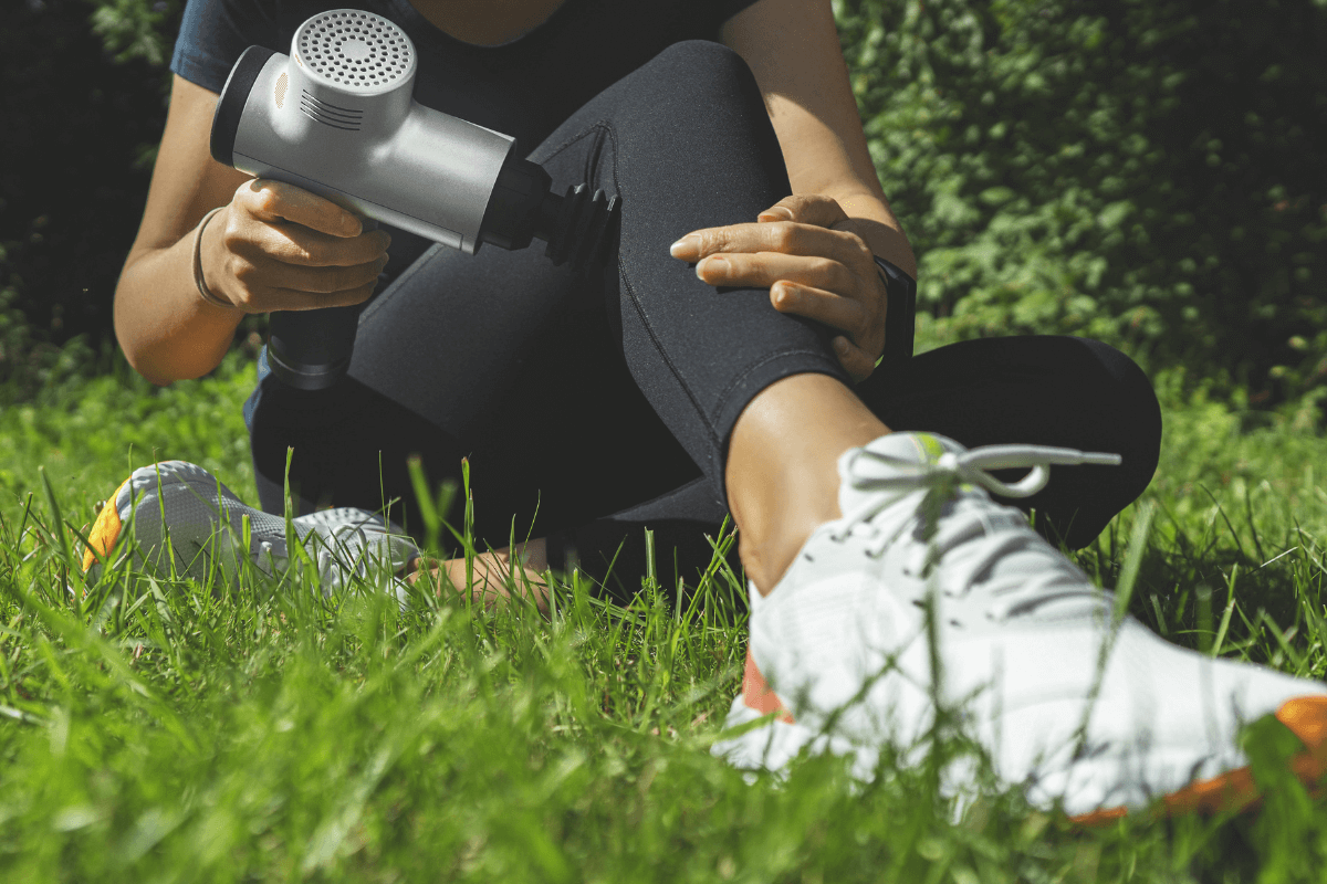 Mulher sentada no gramado demonstrando como usar a pistola massageadora na perna, aplicando o aparelho prateado na região lateral da panturrilha para recuperação muscular.