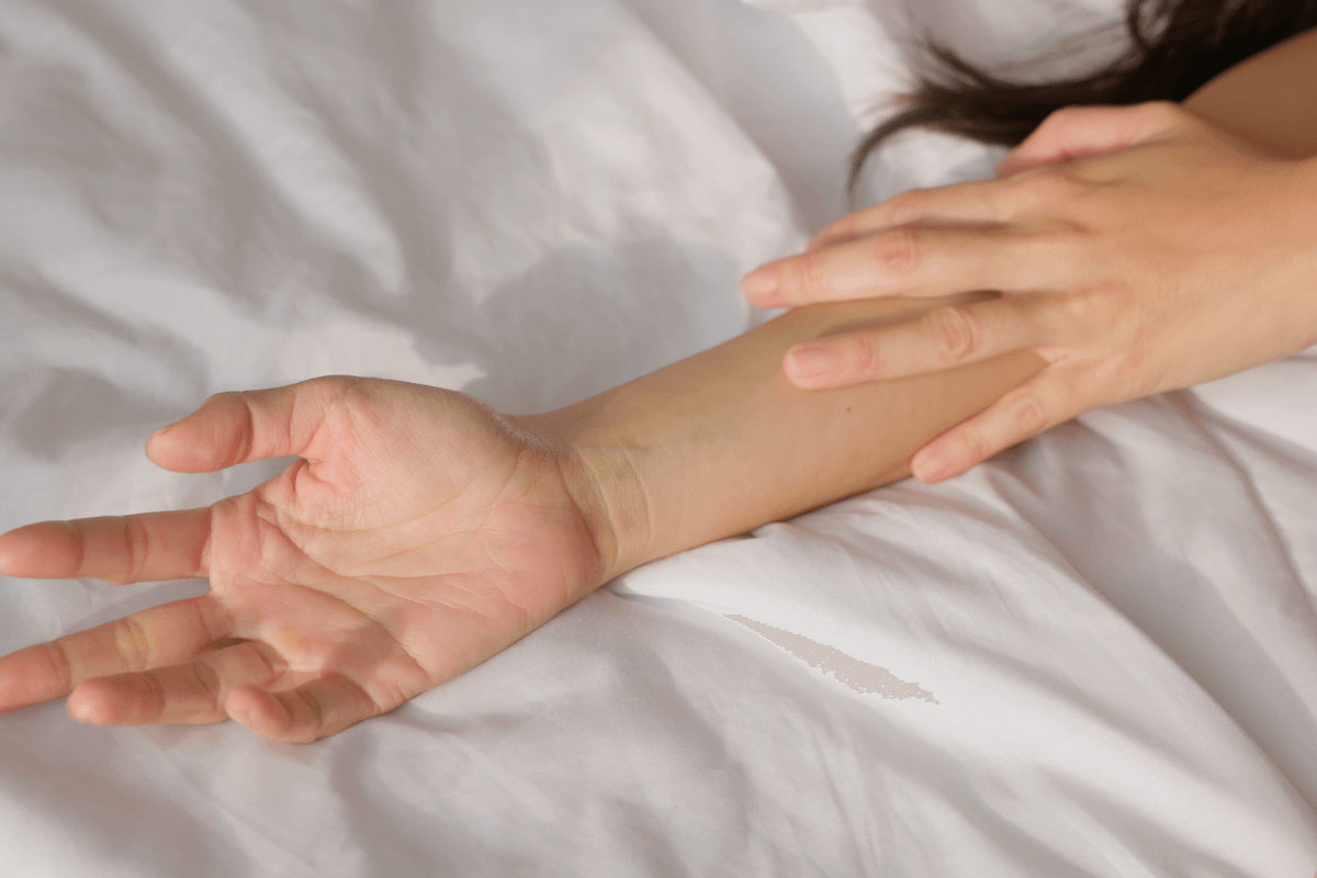 Close de uma mão acariciando suavemente o braço da parceira sobre lençóis brancos, representando a comunicação sem palavras e a sensibilidade ao toque.