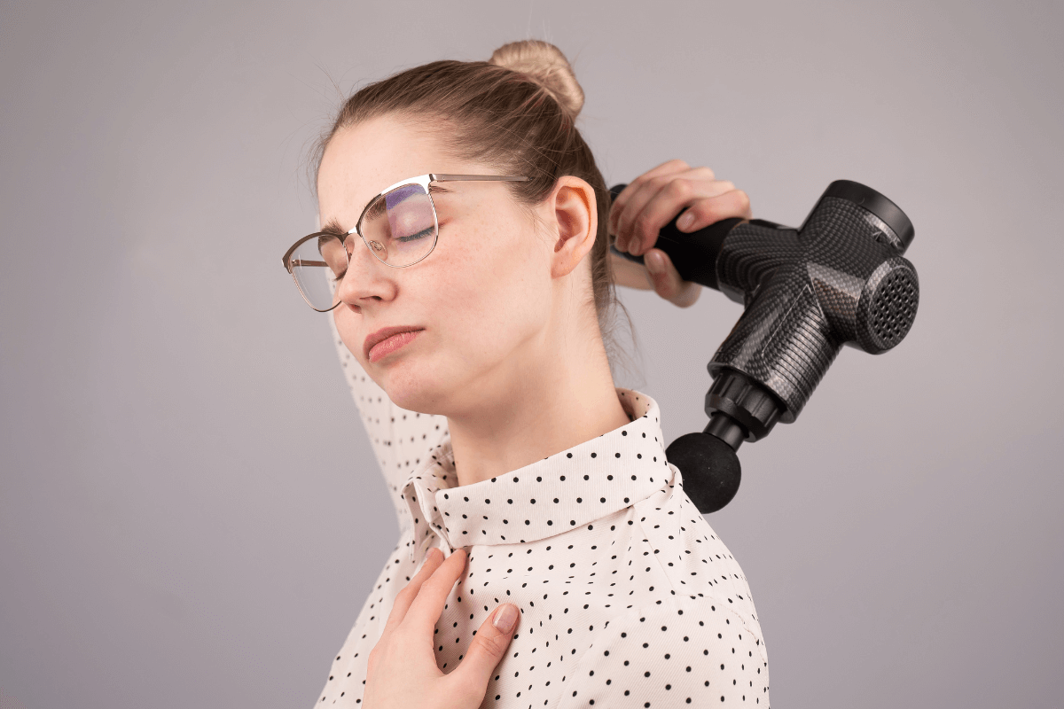 Dor terapêutica: Identificando o limite do relaxamento Mulher aplicando pistola de massagem no músculo trapézio com expressão de alívio concentrado, ilustrando a sensação de 'dor terapêutica' ou dor boa durante a liberação miofascial.