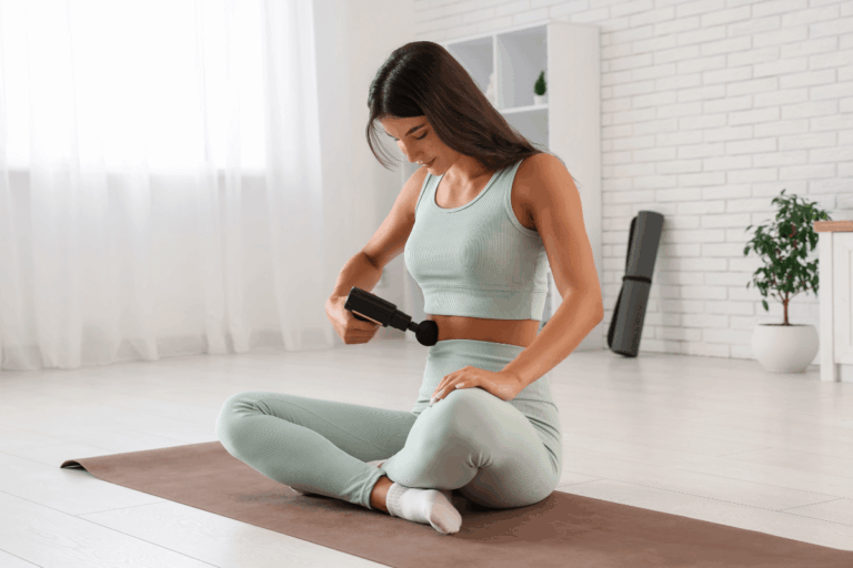 Mulher sentada em tapete de yoga aplicando pistola massageadora suavemente na região lateral do abdômen, demonstrando como é bom usar massageador na barriga para relaxamento e alívio.