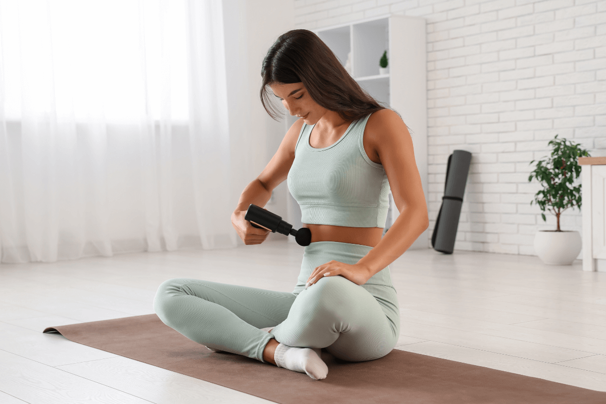 Mulher sentada em tapete de yoga aplicando pistola massageadora suavemente na região lateral do abdômen, demonstrando como é bom usar massageador na barriga para relaxamento e alívio.