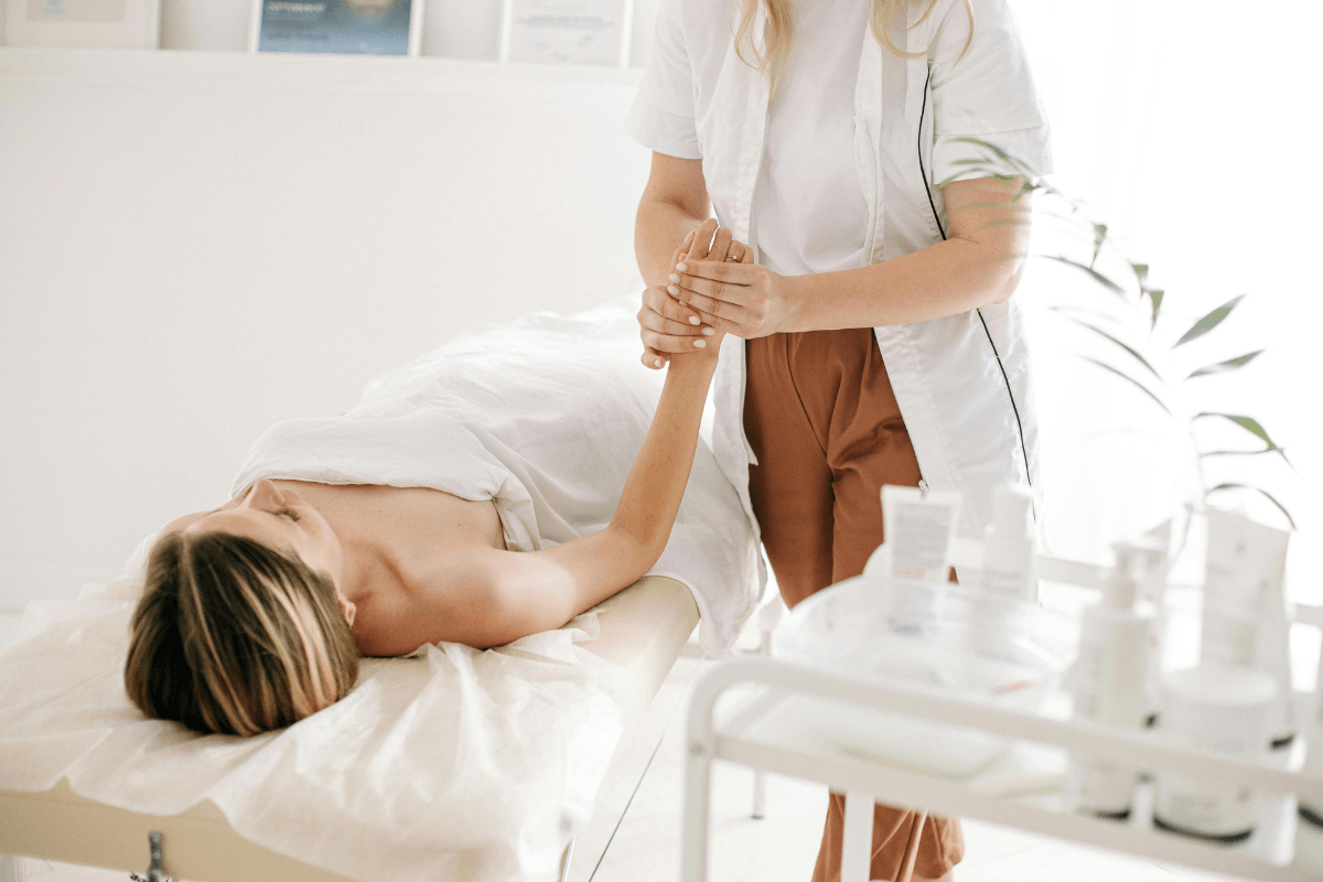 Biossegurança na massagem: Uniforme, lençóis limpos e ambiente organizado Massagista profissional usando uniforme branco limpo realizando atendimento em maca com lençóis estéreis, exemplificando a higiene e biossegurança na sala de atendimento.