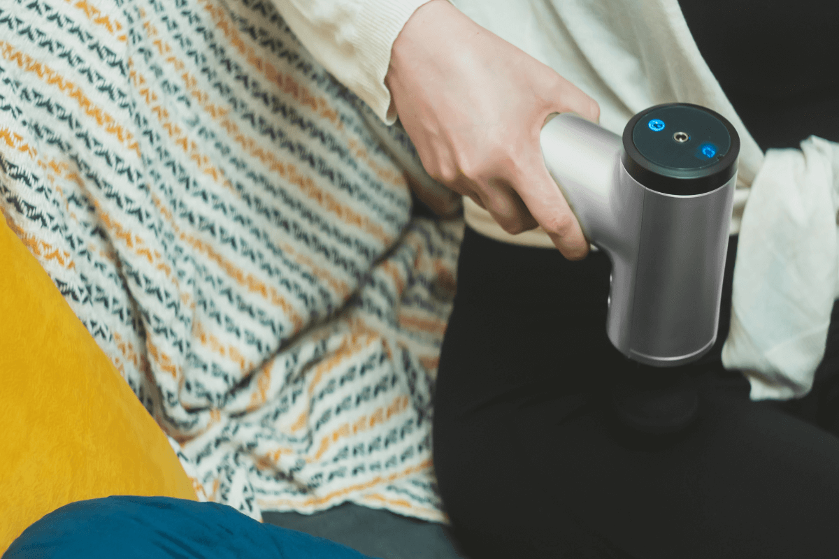 Posição correta para massagem no quadríceps Pessoa sentada aplicando a pistola massageadora na frente da coxa para soltar o músculo quadríceps com movimento vertical.