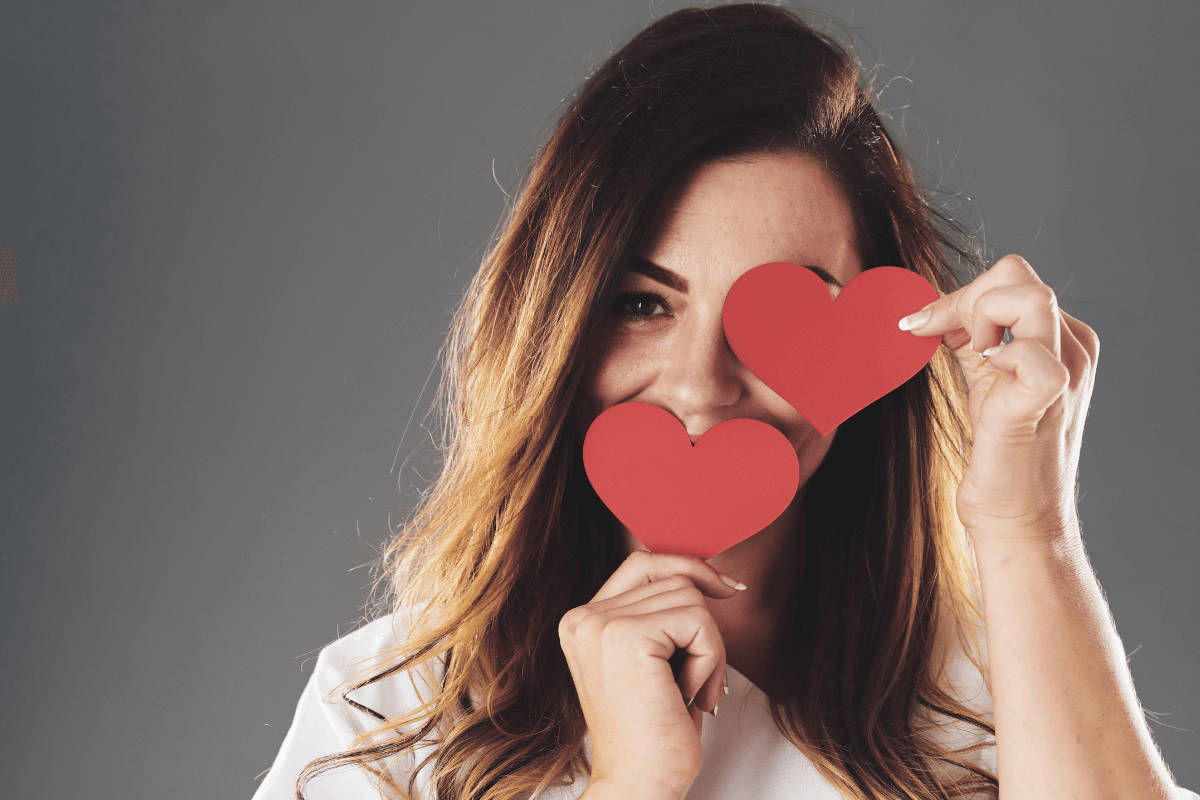 O poder da curiosidade e do mistério para manter o interesse Mulher sorridente cobrindo os olhos com dois corações de papel vermelho, simbolizando a importância de não revelar tudo de uma vez e manter o mistério na interação romântica.