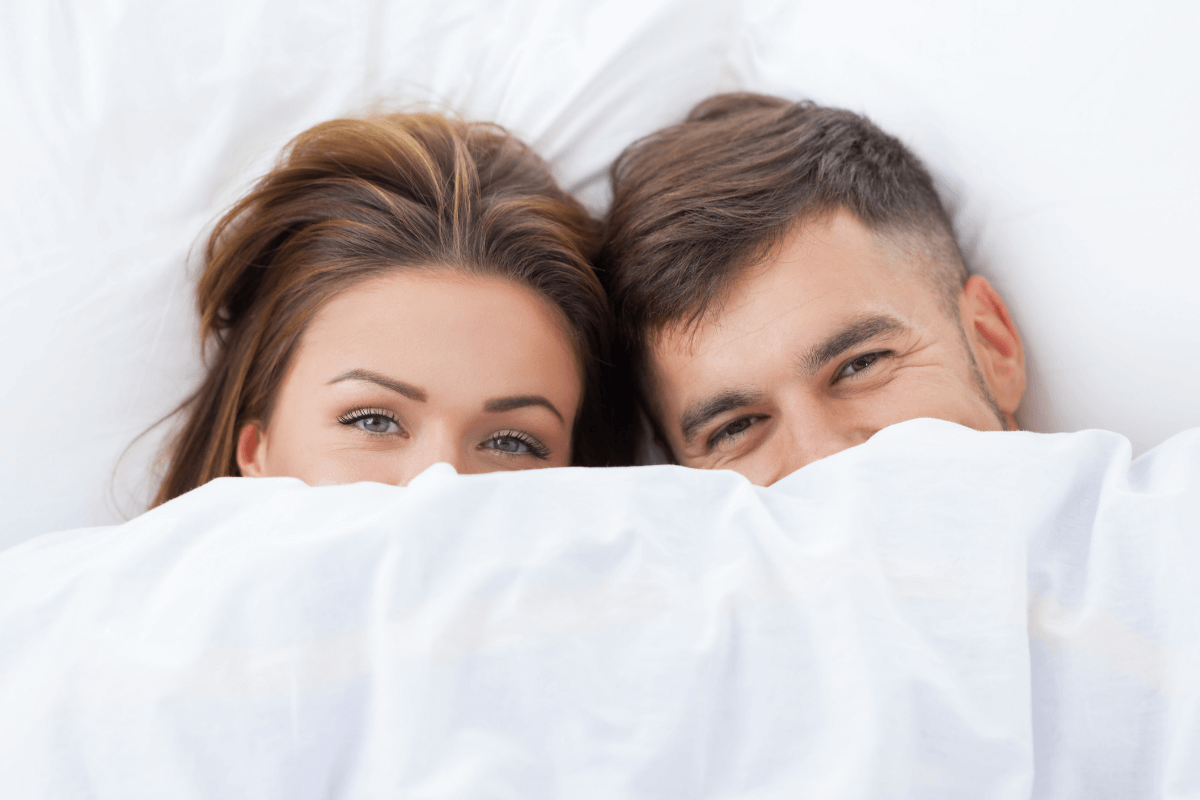 O silêncio estratégico: Deixando o olhar falar mais alto Casal na cama com apenas os olhos visíveis sob o lençol, ilustrando o silêncio estratégico e a comunicação intensa pelo olhar após o dirty talk.