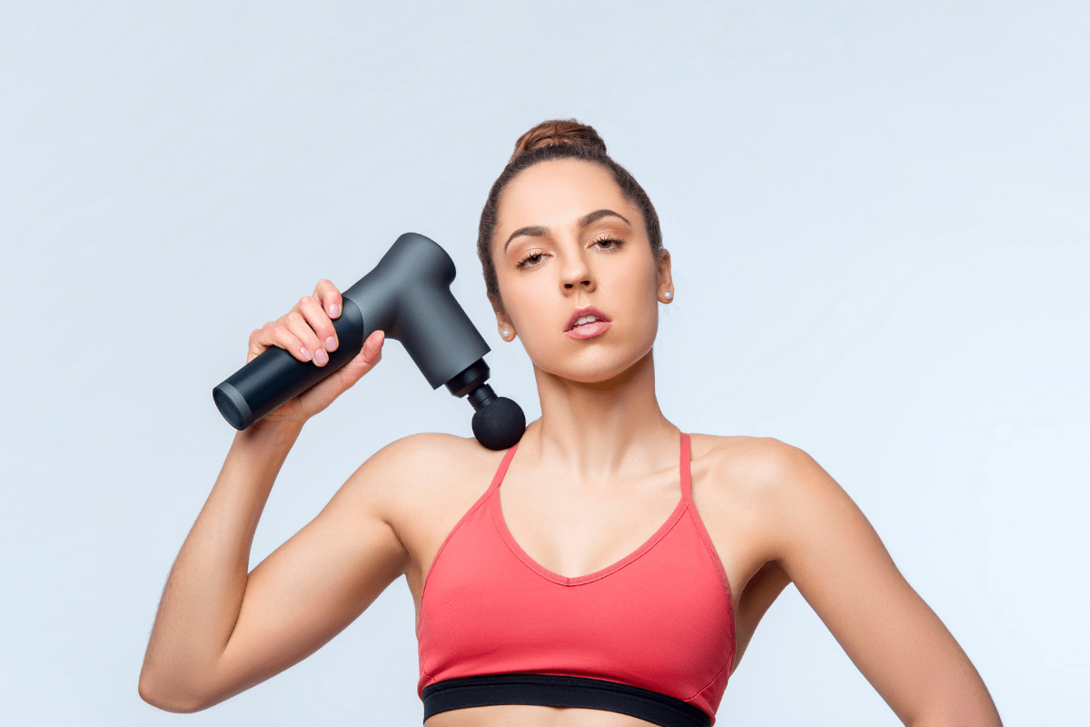 Mulher segurando uma pistola massageadora próxima ao ombro e pescoço, ilustrando o artigo sobre onde não se pode colocar o massageador elétrico para evitar lesões em áreas sensíveis e perigosas.