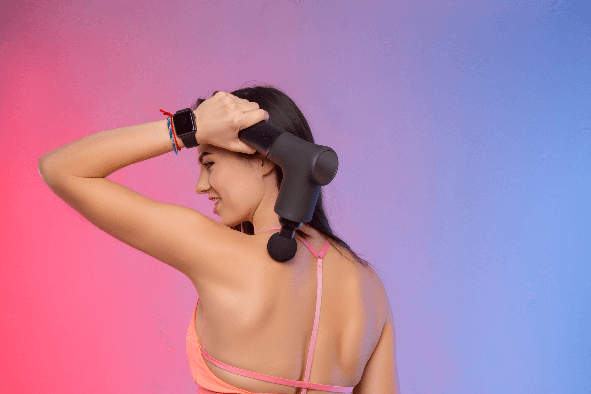 Mulher aplicando massageador no músculo do trapézio, exemplificando o cuidado de evitar estruturas ósseas e articulações expostas como a coluna vertebral e as escápulas durante o uso.