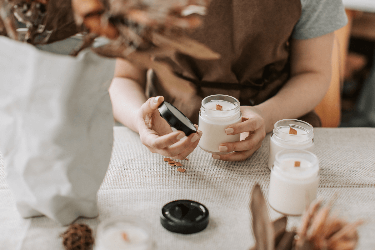 Mãos segurando delicadamente uma vela vegetal aromática com pavio de madeira em pote de vidro, ilustrando para que serve passar vela no corpo para hidratação e relaxamento muscular.