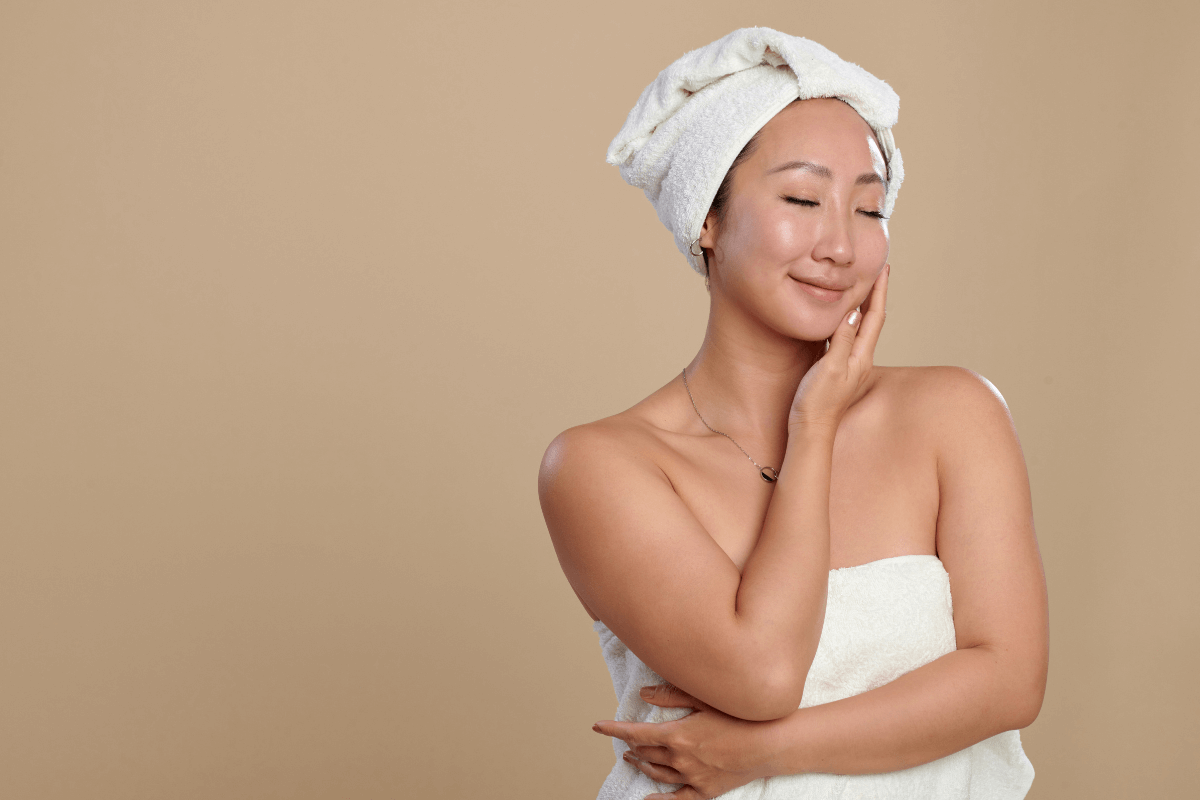 Mulher asiática sorrindo com toalha de banho na cabeça e no corpo, tocando o rosto e exibindo a pele do ombro com aparência úmida e macia.