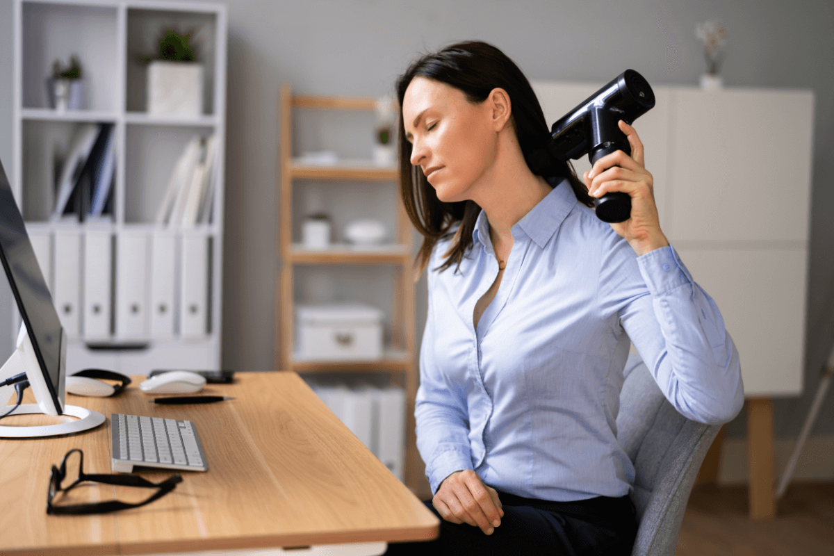 Alívio de tensão no escritório com pistola massageadora. Mulher utilizando pistola massageadora no pescoço durante o expediente no escritório para aliviar a tensão muscular causada pela má postura no computador.