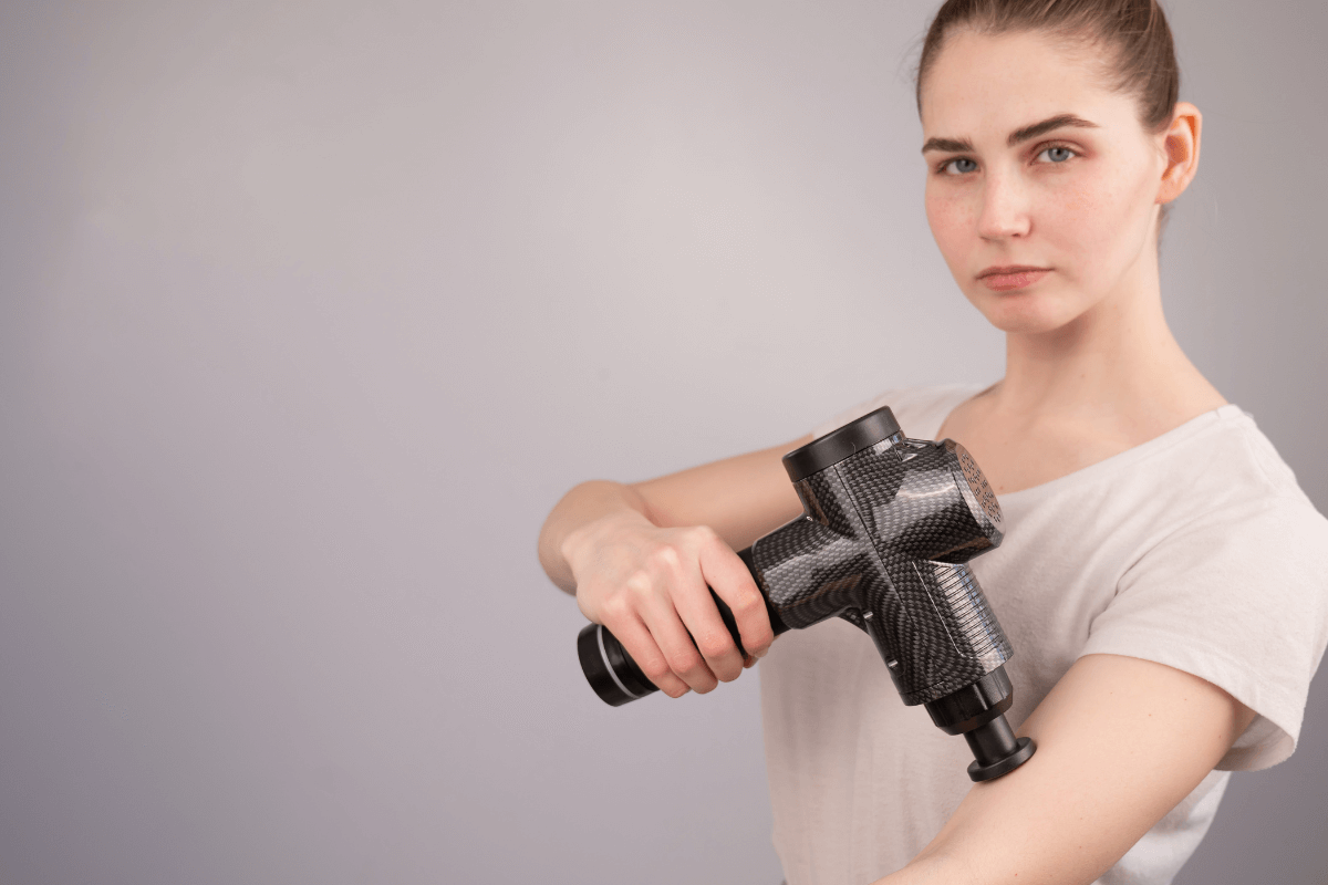 Rotina de relaxamento noturno com pistola massageadora. Mulher realizando automassagem suave no braço com a pistola massageadora para acalmar o sistema nervoso e baixar o nível de cortisol antes de dormir.