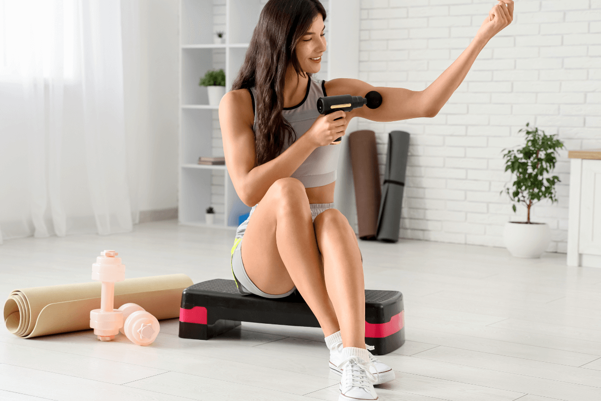 Mulher sentada em equipamento de ginástica aplicando uma pistola de massagem no braço esquerdo, ilustrando a rotina de cuidados para quem busca saber se pode usar massageador todos os dias.