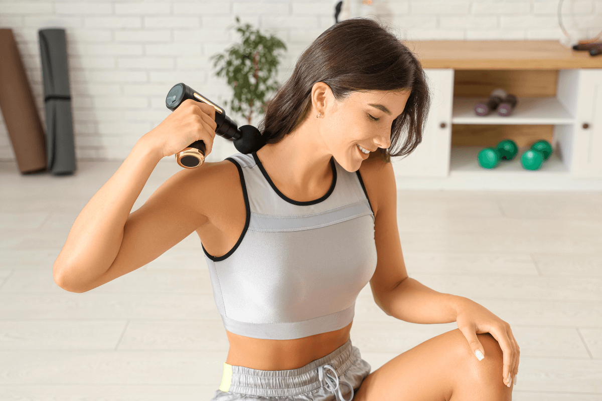 Mulher fitness sorrindo enquanto aplica a pistola massageadora no trapézio para aliviar a tensão e demonstrar na prática qual o benefício do relaxamento muscular imediato.