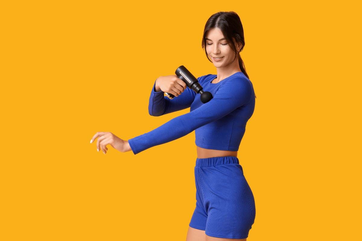 Mulher com roupa esportiva azul aplicando pistola massageadora no braço em fundo amarelo, ilustrando o guia sobre quantos minutos devo usar o massageador elétrico com segurança.