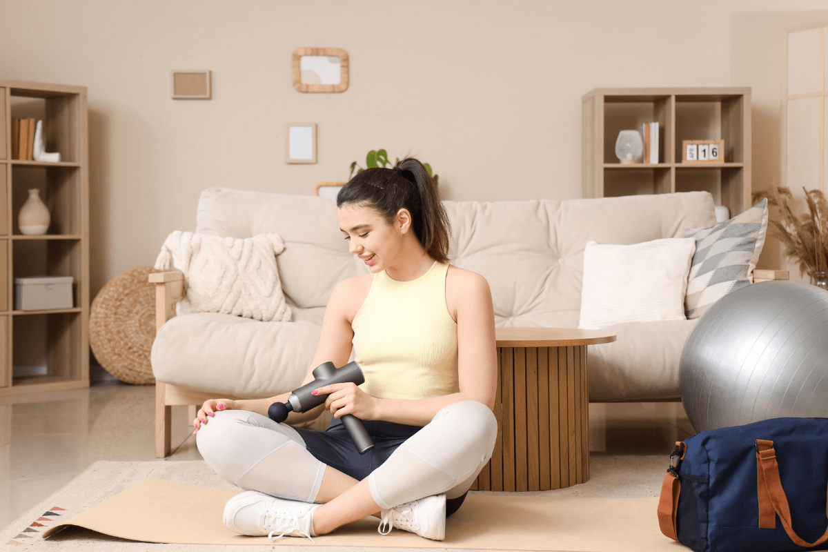 Alívio muscular através da circulação e relaxamento Mulher sorrindo sentada no tapete de yoga segurando uma pistola massageadora, representando o alívio muscular e a melhora da circulação pós-treino, desmistificando a quebra de ácido lático.