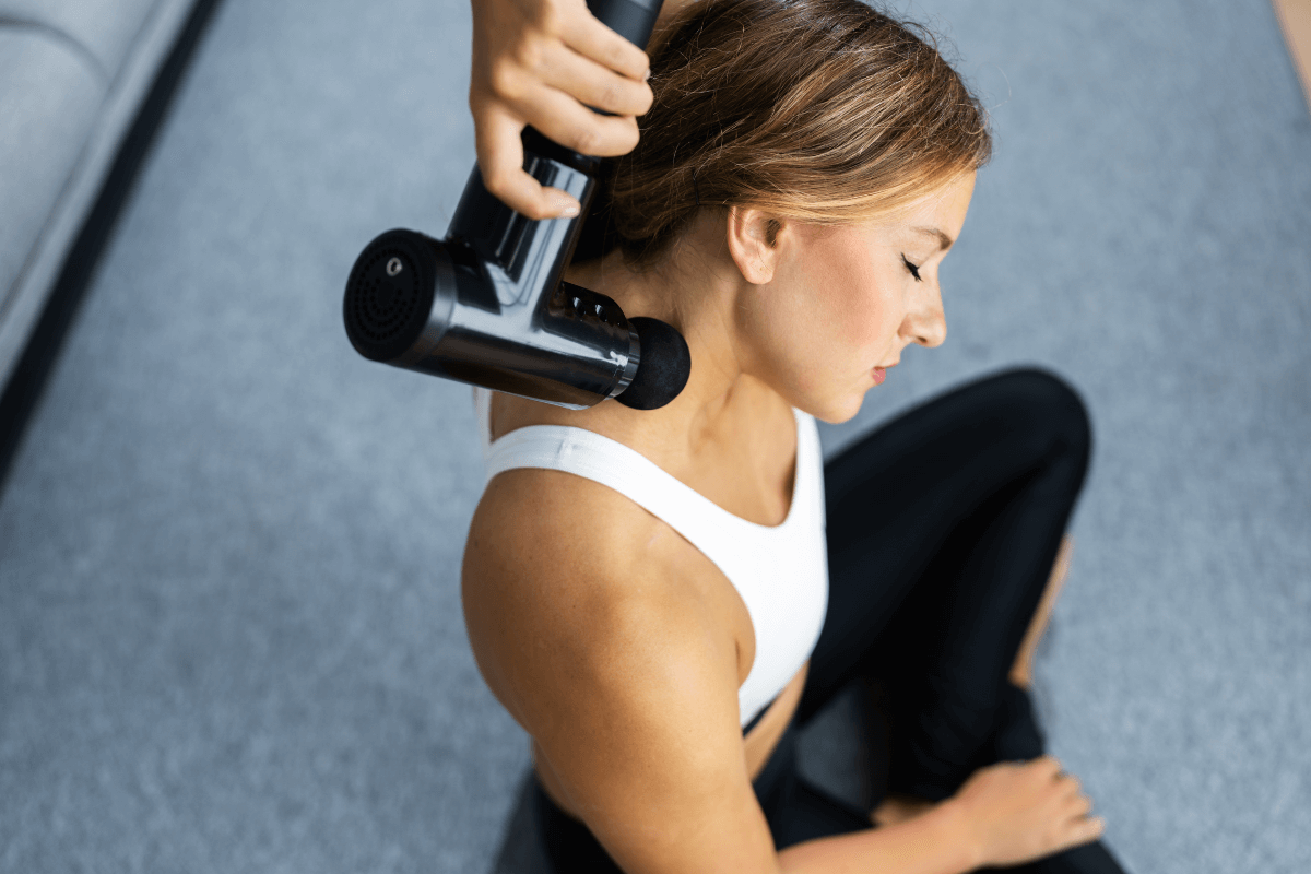 Mulher aplicando pistola massageadora na região do trapézio e pescoço, uma área sensível onde o risco da superestimulação por tempo excessivo pode causar desconforto.