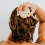 Mulher tomando banho de costas lavando o cabelo, ilustrando a rotina de uso de óleos corporais no chuveiro.
