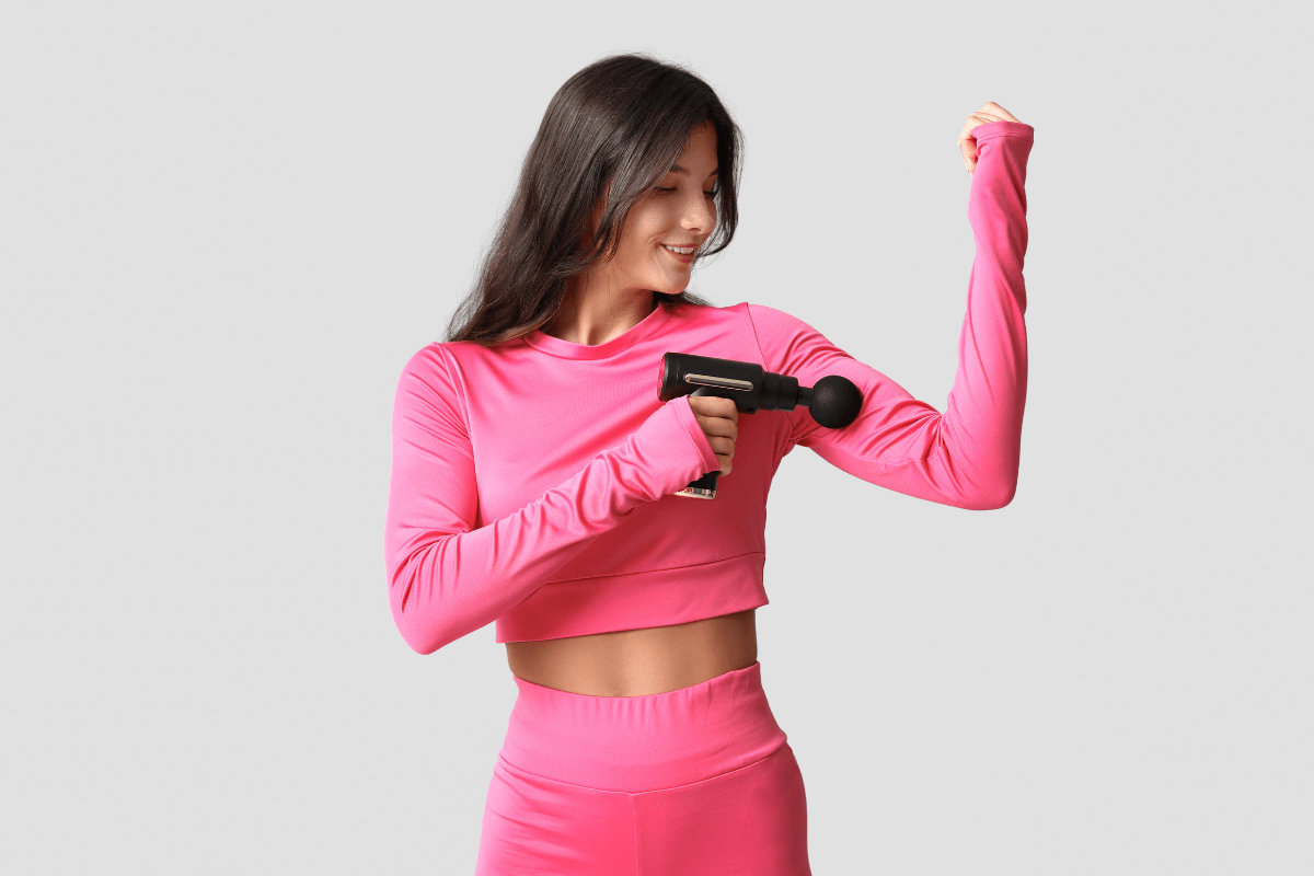 Mulher vestindo roupa rosa fitness aplicando massageador tipo pistola no bíceps esquerdo, demonstrando o foco em um grupo muscular por vez para respeitar o tempo de uso seguro.