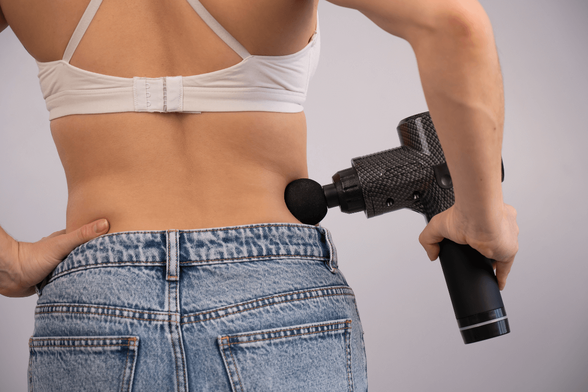 Massagem na lombar: Onde aplicar sem atingir os rins Mulher aplicando pistola de massagem na musculatura lateral da lombar (quadrado lombar), demonstrando a distância segura da coluna vertebral e evitando a percussão direta sobre a área dos rins.